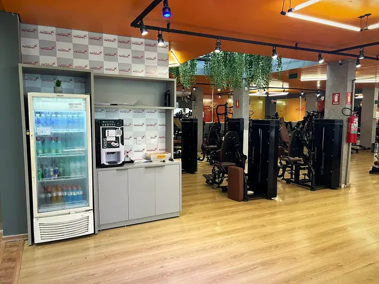 Geladeira de bebidas e máquina de café instaladas dentro do salão, ao lado das estações de musculação.