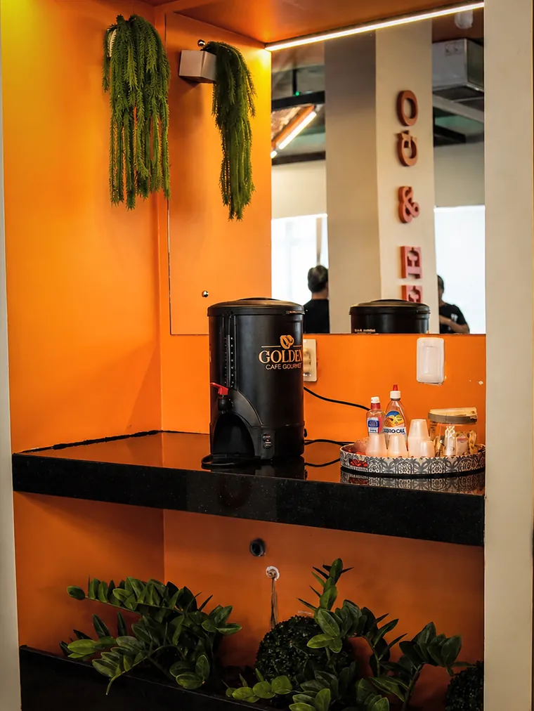 Nicho laranja com bancada de granito preto, a garrafa térmica de café Golden, copinhos, adoçante e plantas penduradas.