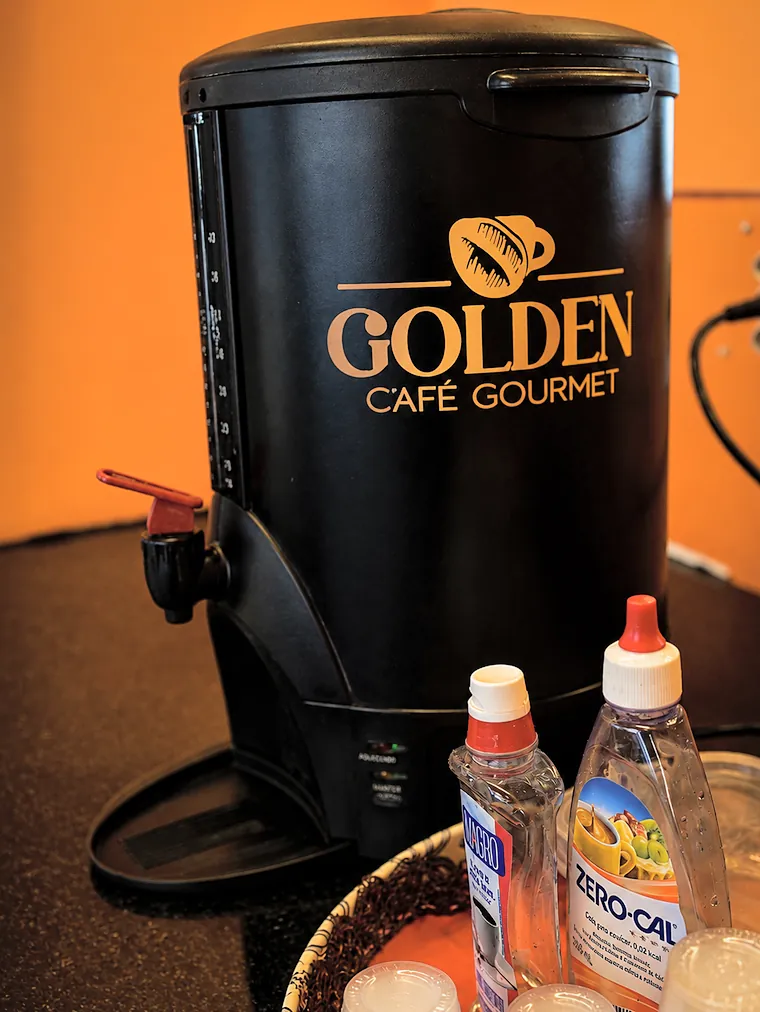 Garrafa térmica preta de café com a marca Golden Café Gourmet, ao lado de frascos de adoçante.