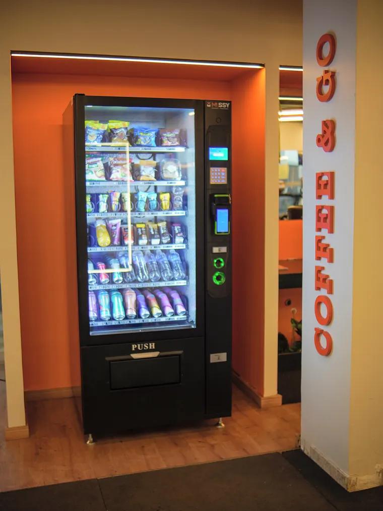 Máquina de lanches e bebidas iluminada num nicho laranja, ao lado da coluna com as letras Coffee & Go.