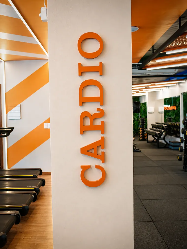 Coluna branca com a palavra Cardio em letras laranja, em pé, com esteiras ao fundo.