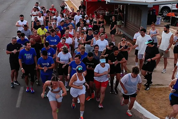 O grupo do clube saindo em corrida pela avenida, visto de cima pelo drone, com as tendas vermelhas ao fundo.