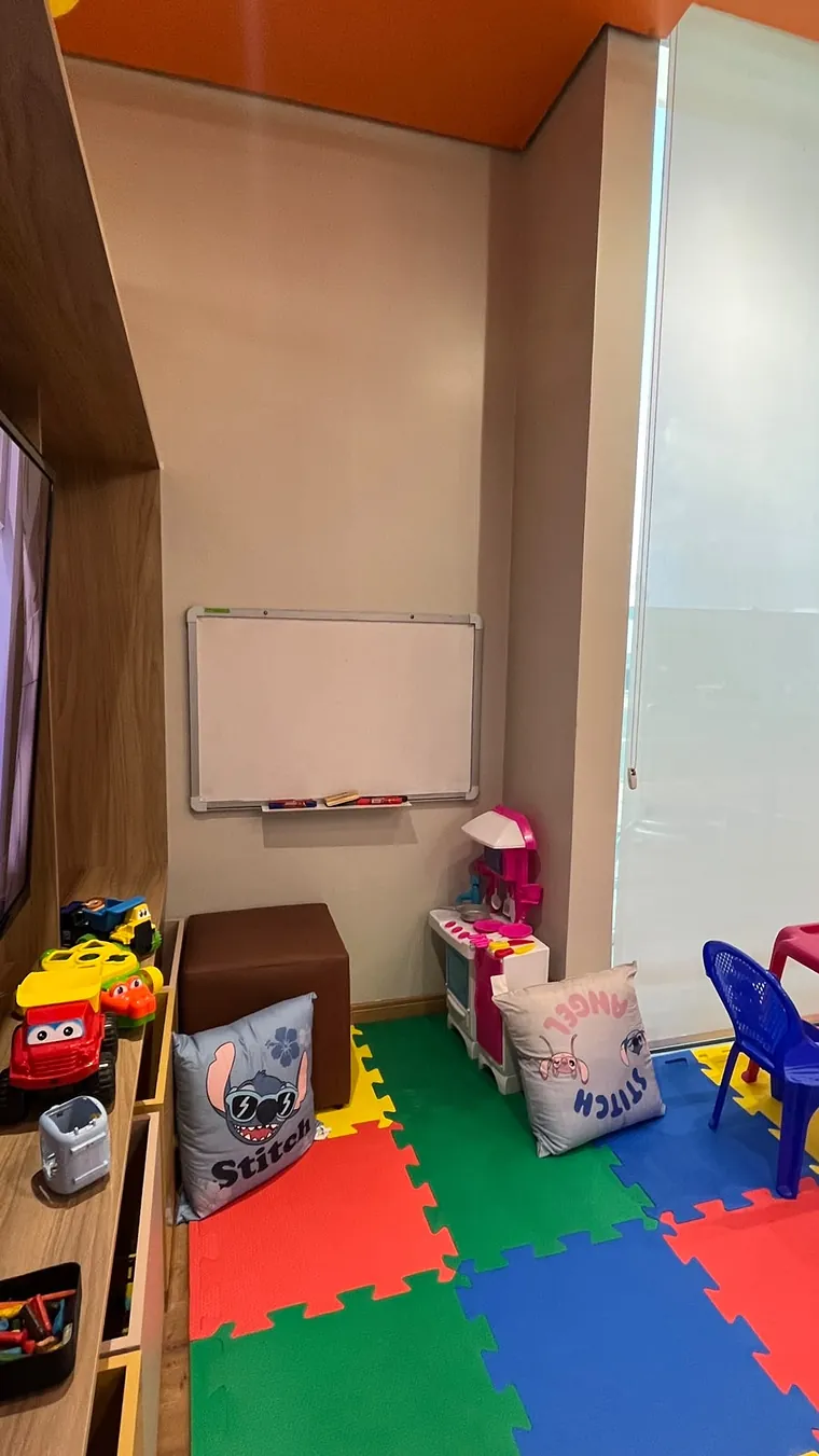 Lousa branca na parede, pufe marrom, carrinho de brinquedo, cozinha de brinquedo e almofadas sobre o piso de EVA.
