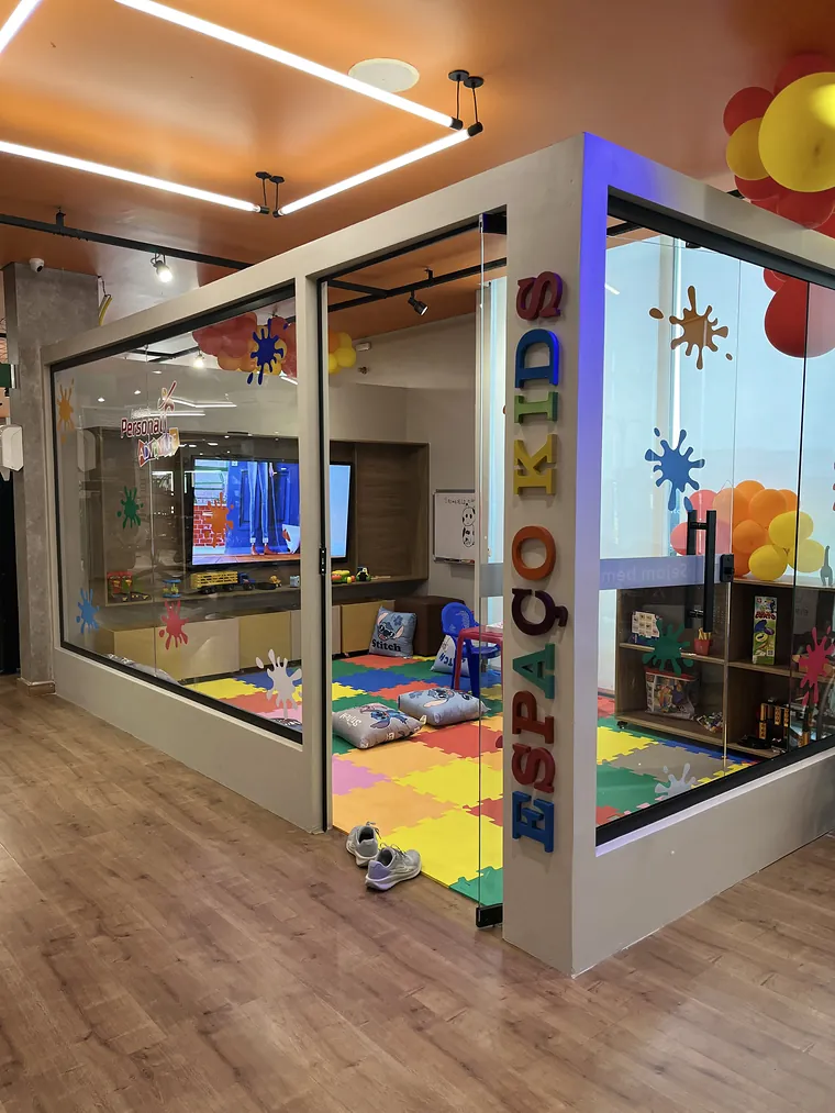 A sala do Espaço Kids, de paredes de vidro, com as letras Espaço Kids em pé na coluna da quina, piso de EVA colorido, almofadas e a TV ao fundo.