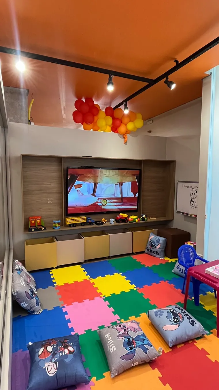 A parede da TV com painel de madeira, carrinhos na prateleira, caixotes de brinquedo e almofadas sobre o piso de EVA colorido.