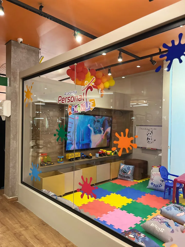 A vitrine de vidro do Espaço Kids com a arte Academia Personal Adventure, manchas de tinta coloridas, a TV e carrinhos na prateleira.
