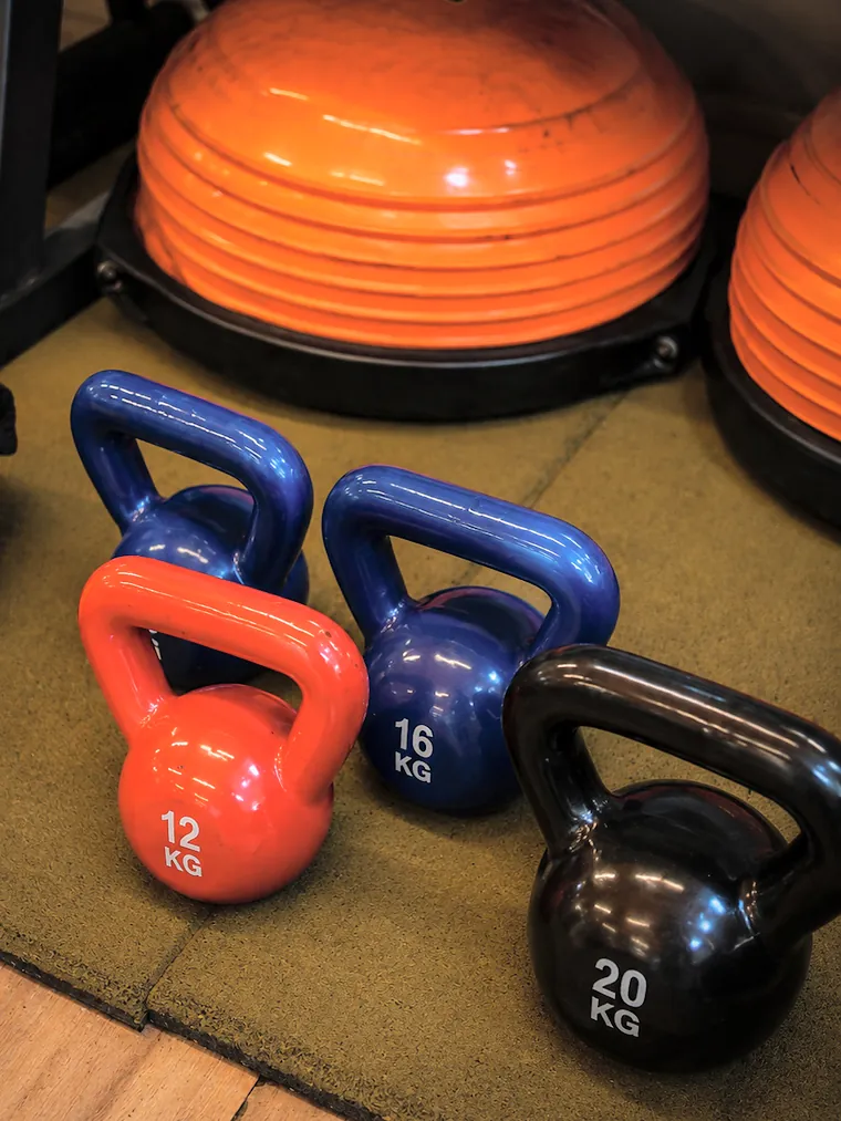 Kettlebell vermelho de 12 quilos, azul de 16 e preto de 20, ao lado de bosus laranja.
