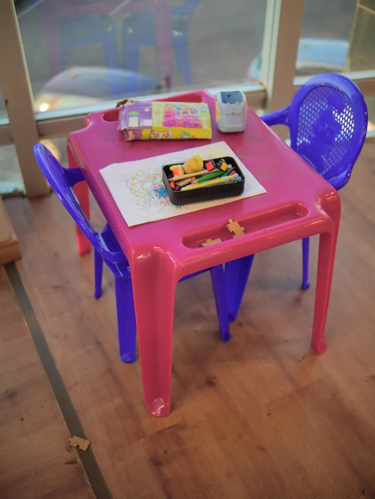 Mesa infantil rosa com cadeiras azuis, lápis de cor e um desenho para colorir, junto à janela.
