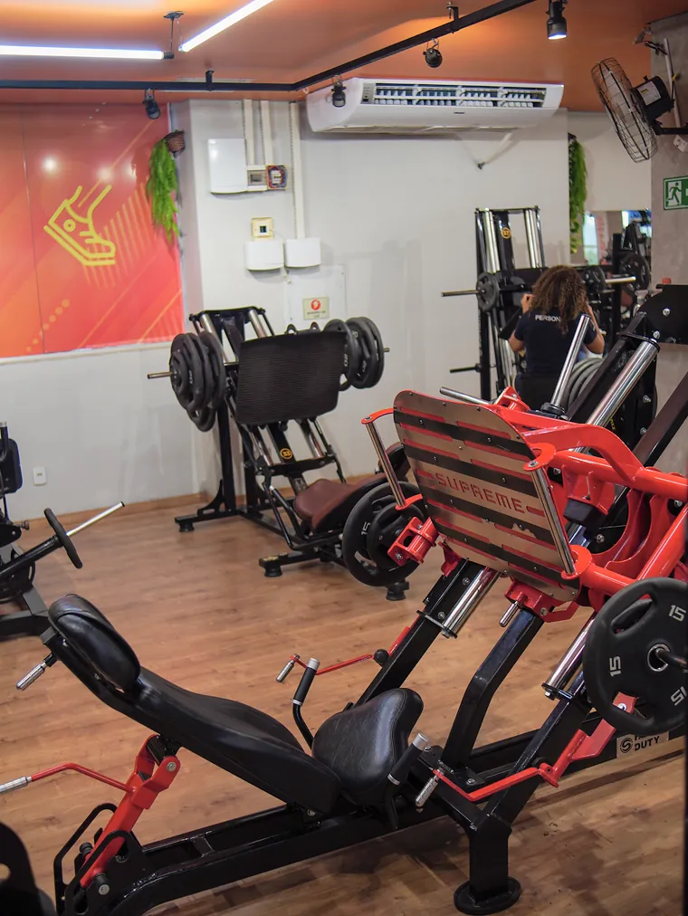 Leg press vermelho e preto carregado com anilhas, diante de um painel laranja na parede.