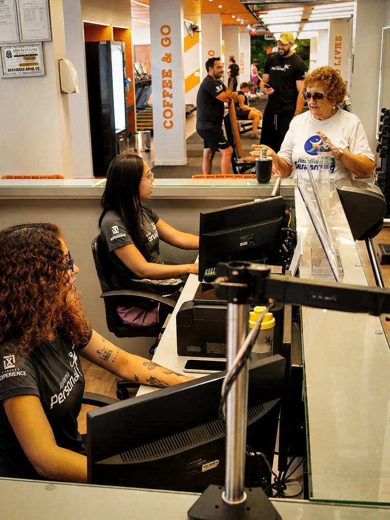 Recepcionistas uniformizadas atendendo uma cliente no balcão, com o salão ao fundo.