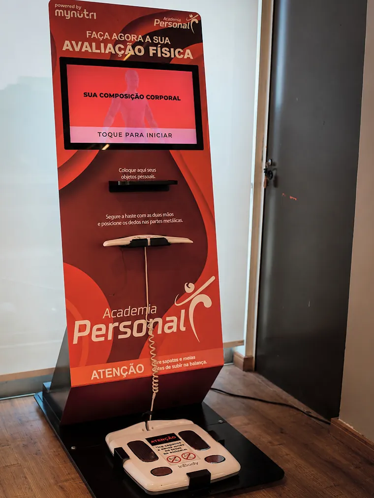 Totem vermelho da Academia Personal com a tela escrita Faça agora a sua avaliação física, a haste para as mãos e a balança no chão.