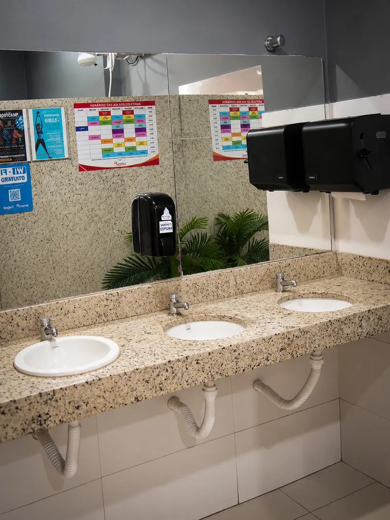 Bancada de granito com três pias, espelho e dispenser de papel.