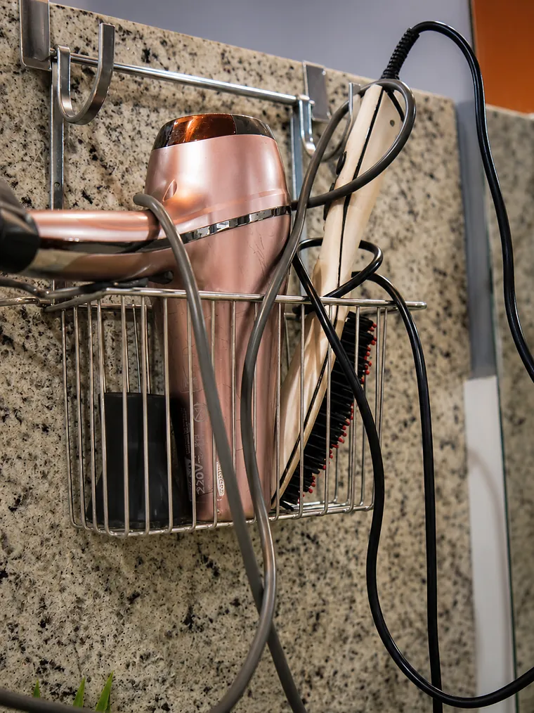 Secador de cabelo rosé e uma prancha alisadora pendurados num suporte na parede de granito.